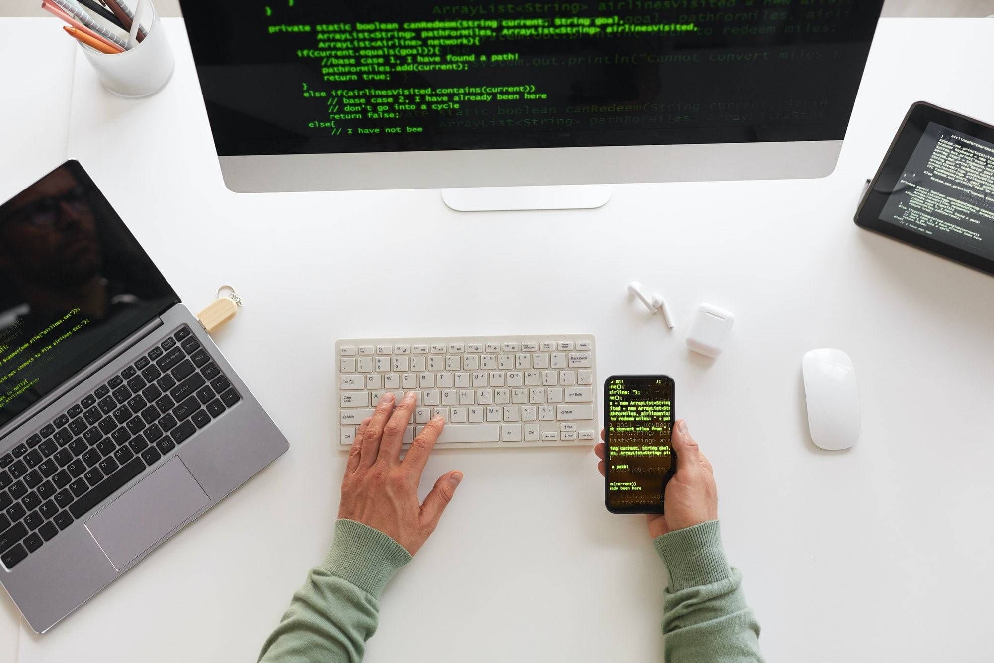 Van bovenaf een bureau met een beeldscherm, laptop, tablet en telefoon die allemaal code tonen