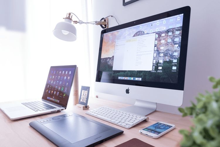 Een iMac en een laptop op een licht bureau, met een tekentablet, een toetsenbord en een telefoon ernaast