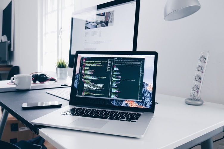 Een laptop met code op het scherm voor een groter beeldscherm met een webpagina, op een licht bureau